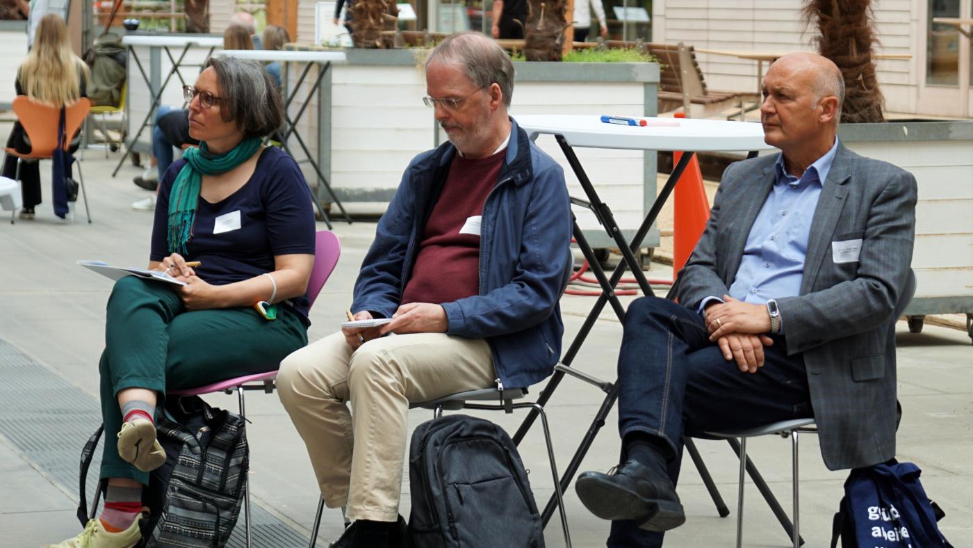 Drei Teilnehmende sitzen auf Stühlen und hören einem Vortrag zu