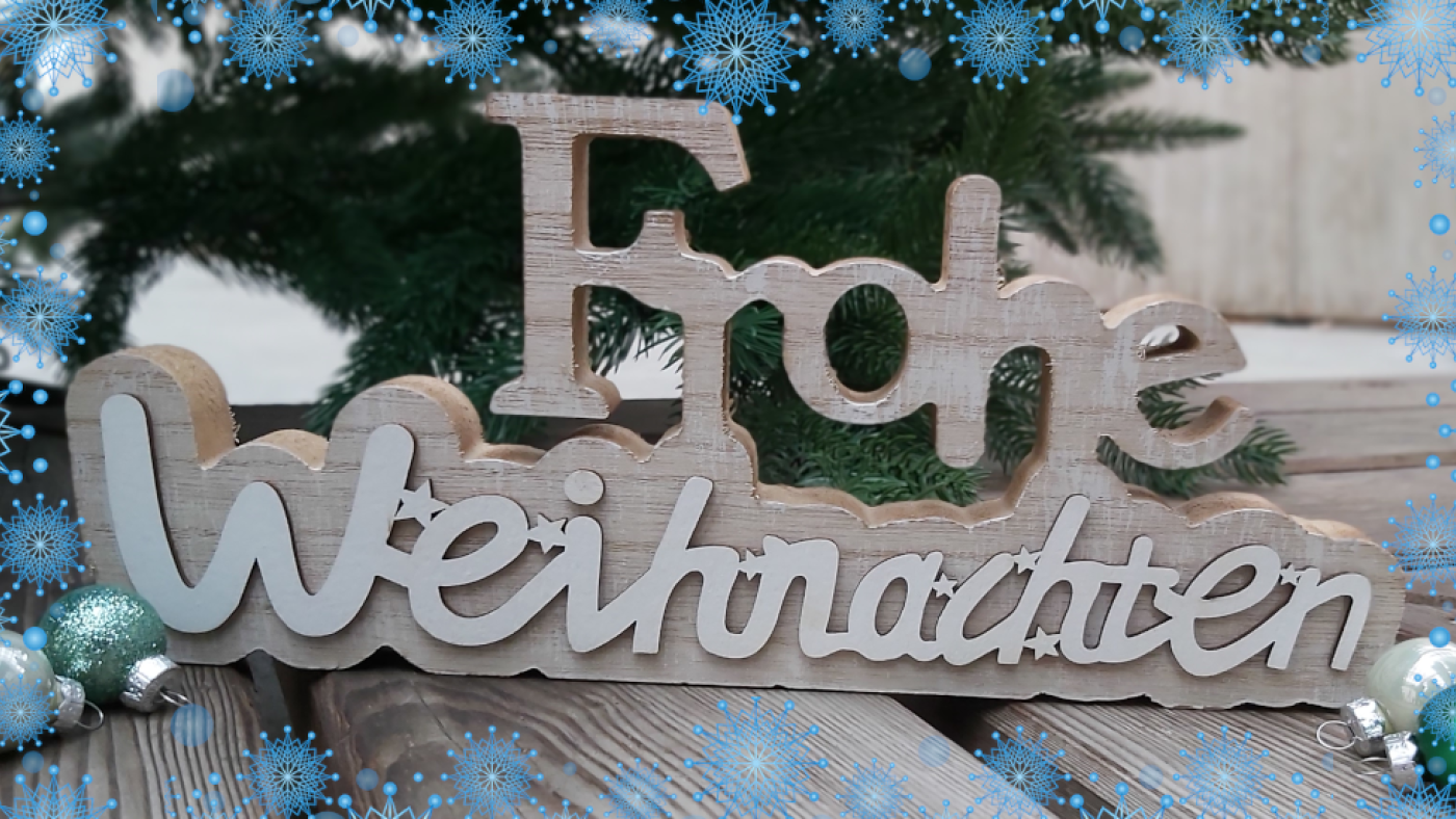 Holzschild "Frohe Weihnachten" steht vor einem Tannenbaum, um das Foto verläuft ein Rahmen aus blauen Schneeflocken