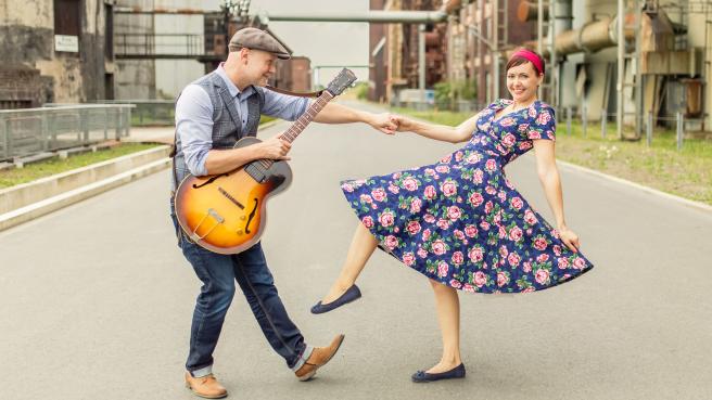 Mann mit Gitarre und Frau tanzen vor Industriekulisse